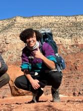 An image of a light skinned biracial Black woman with glasses and curly hair down to her ears. She's posing for a picture in the Grand Canyon with a backpack, a smile, and a thumbs up.